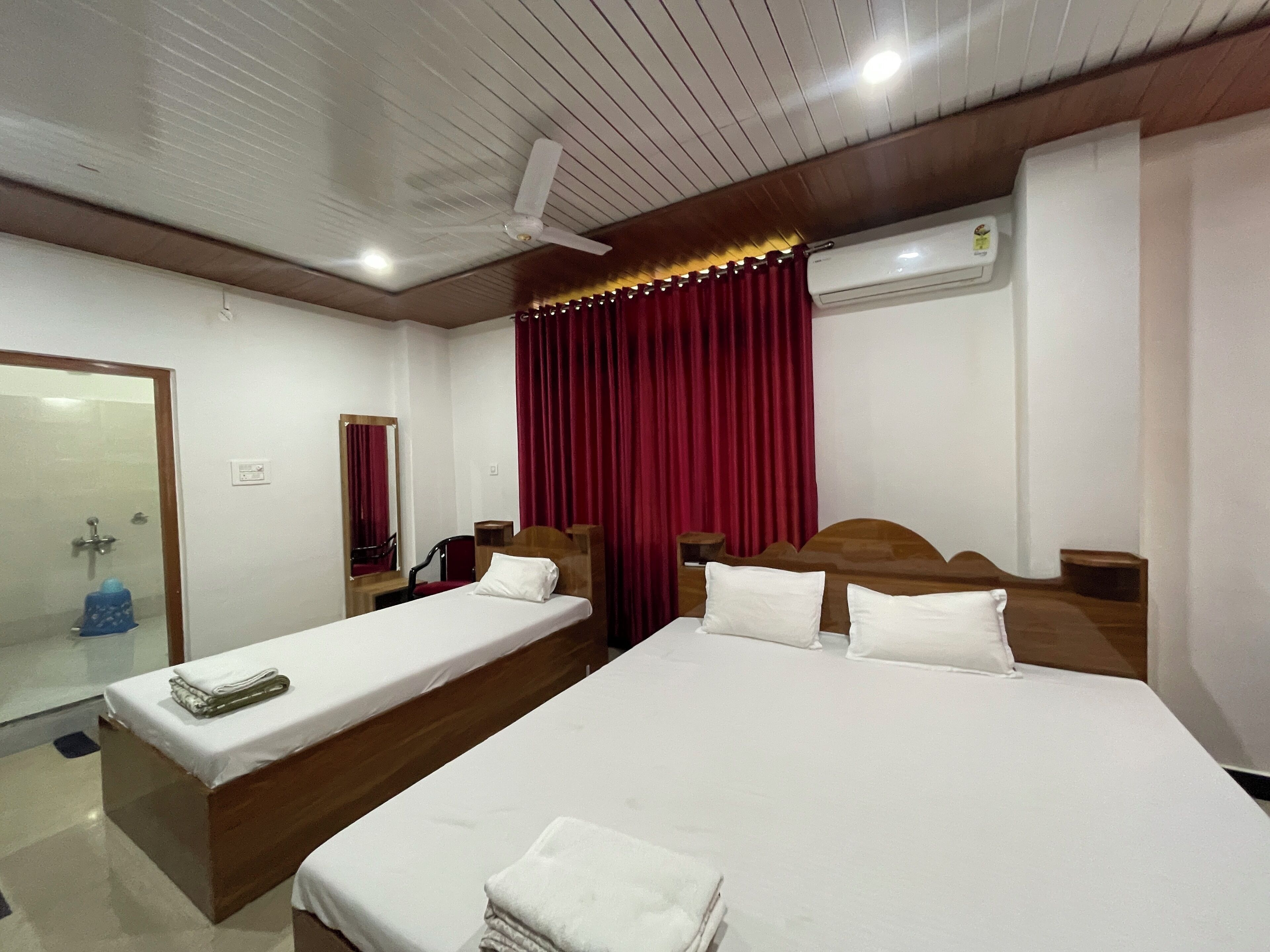 Een hotelkamer met twee bedden, een houten hoofdeinde, een spiegel, een rood gordijn en een airconditioner.
