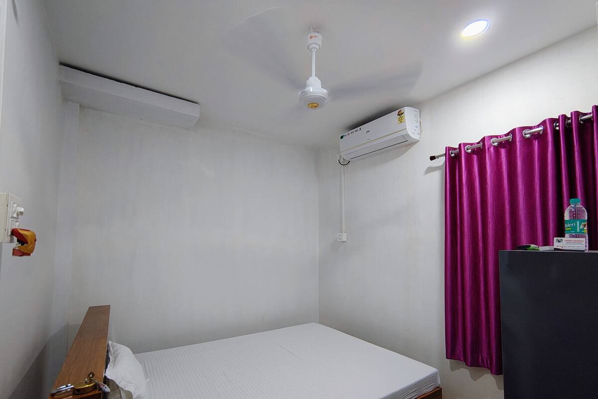 A small, single-bed room with a white ceiling, an air conditioning unit, a ceiling fan, a purple curtain, and a small table with a bottle and a box.