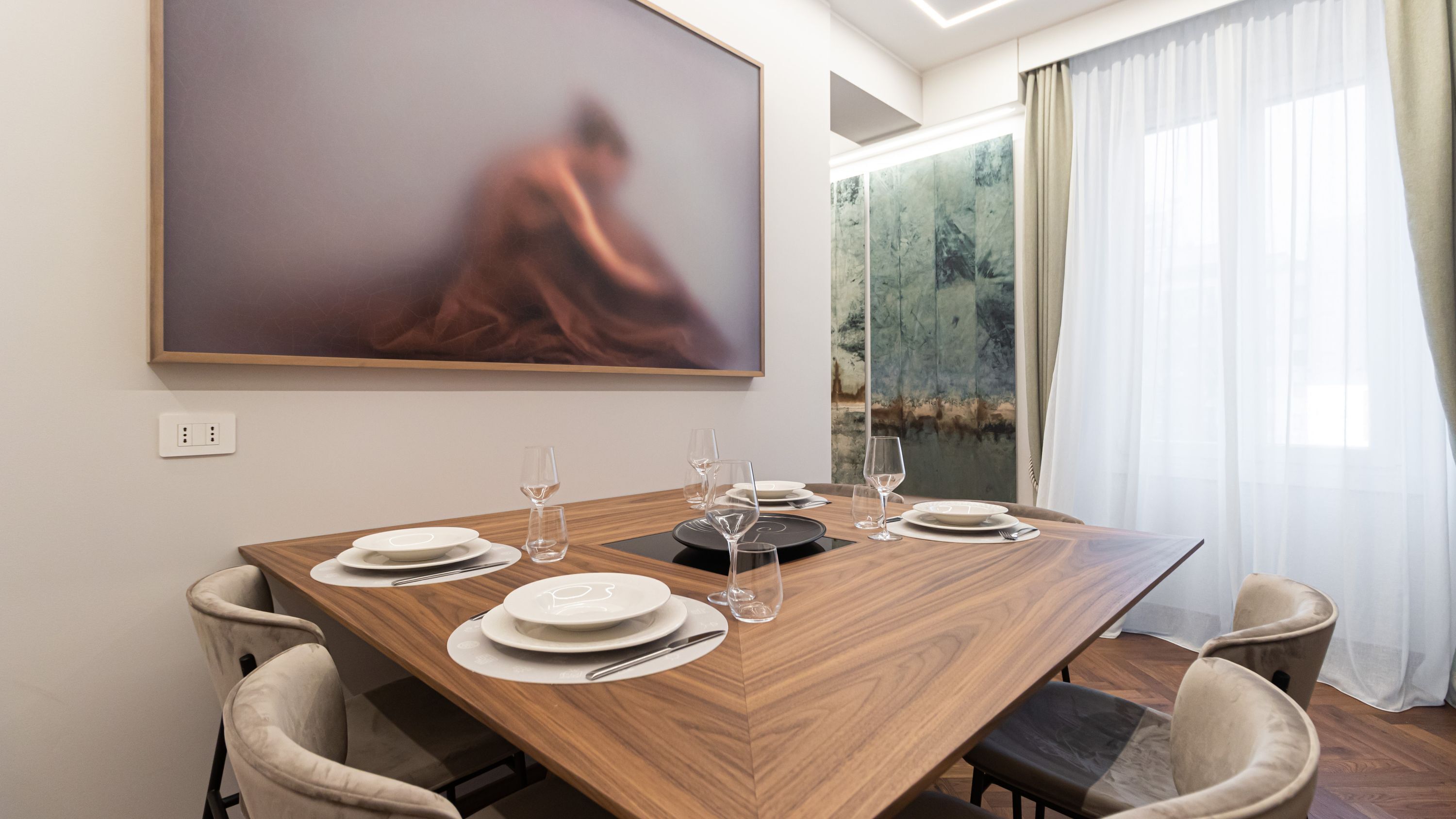 Un comedor moderno con una mesa de madera dispuesta para cuatro personas, rodeada de sillas con cojines. Un cuadro abstracto de gran tamaño adorna la pared, y se aprecia una ventana con cortinas ligeras.