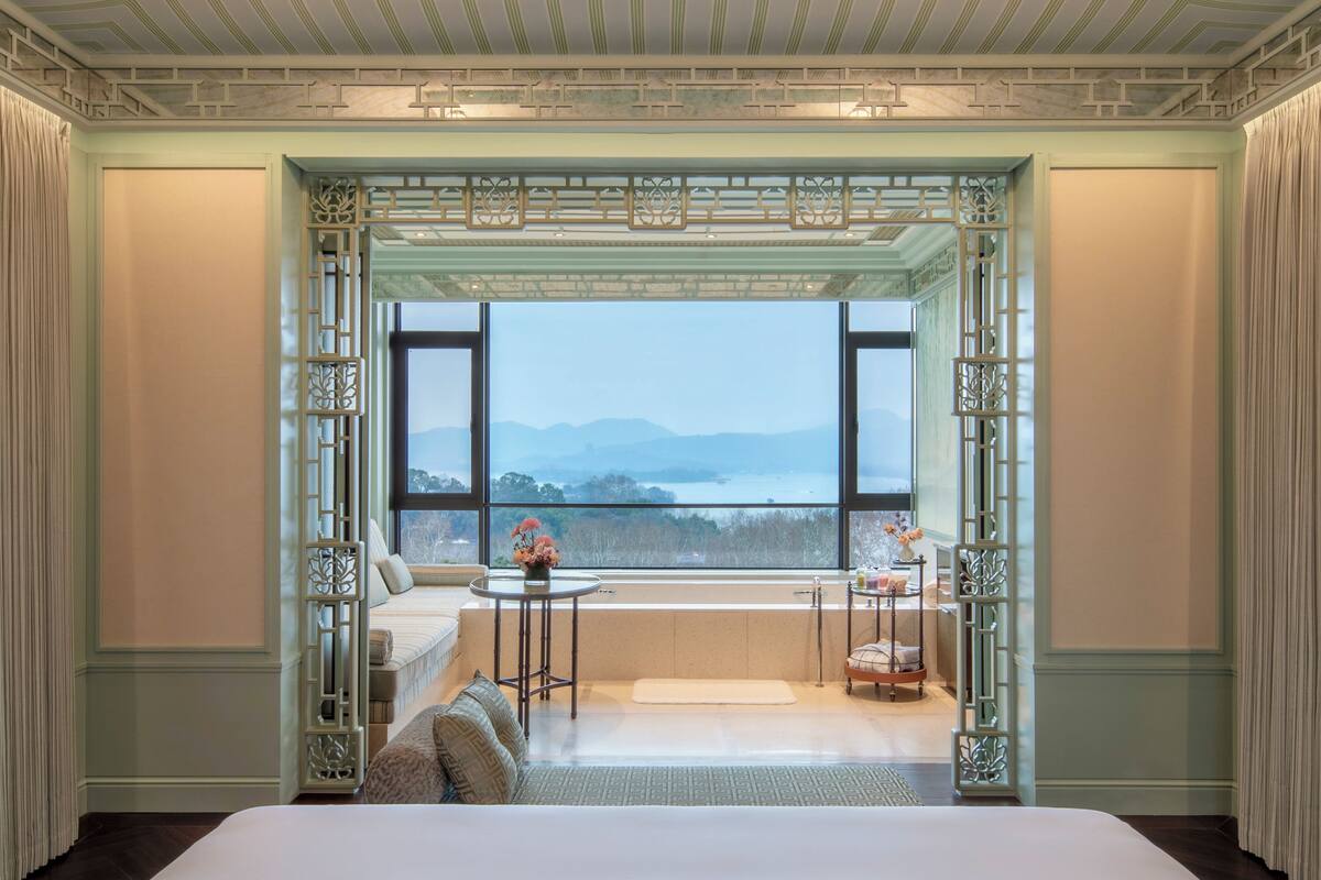 A bedroom with a large window, a bed, a bedside table, and a chair.