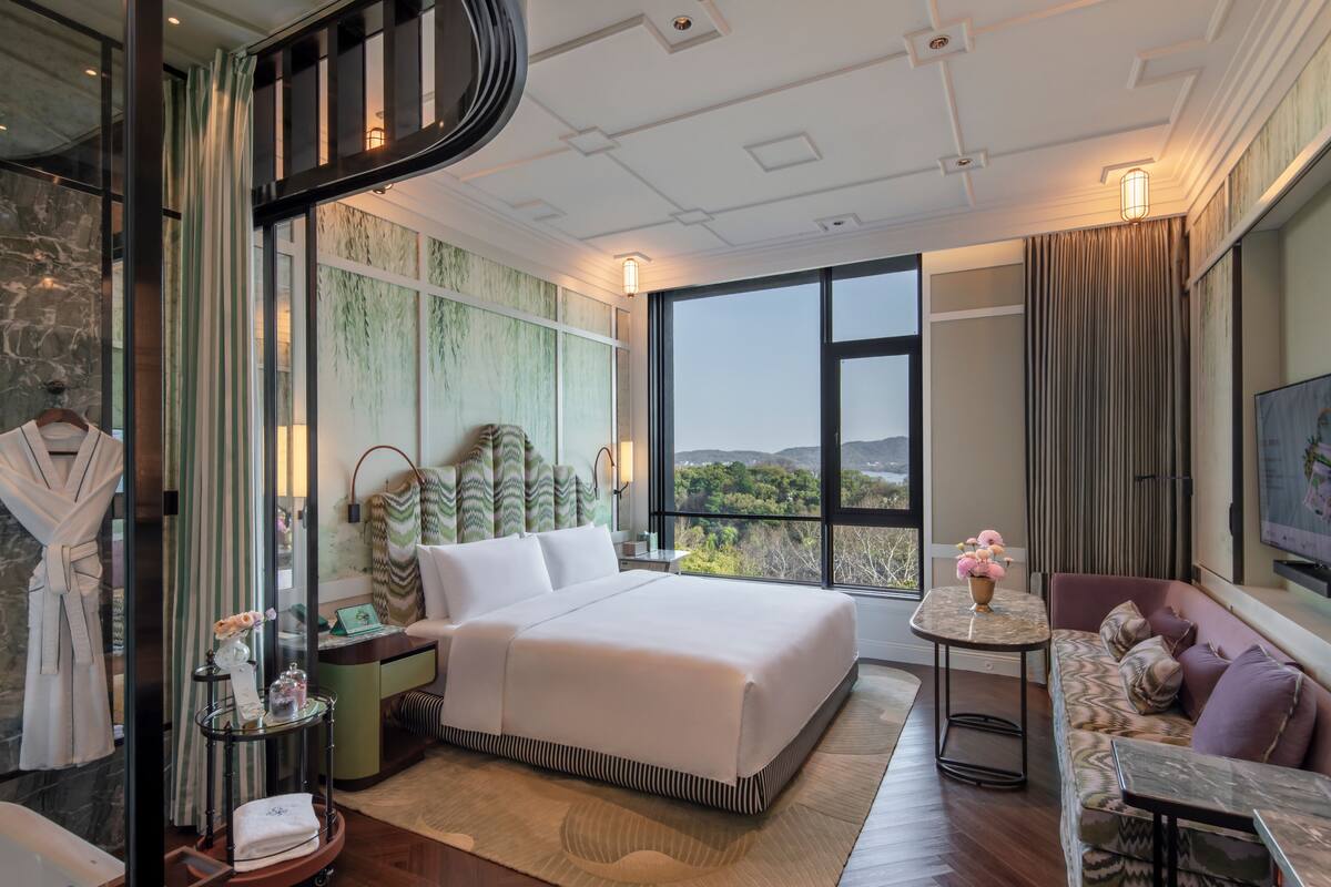 A modern hotel room with a large bed, a sofa, a TV, and a view of the outdoors.