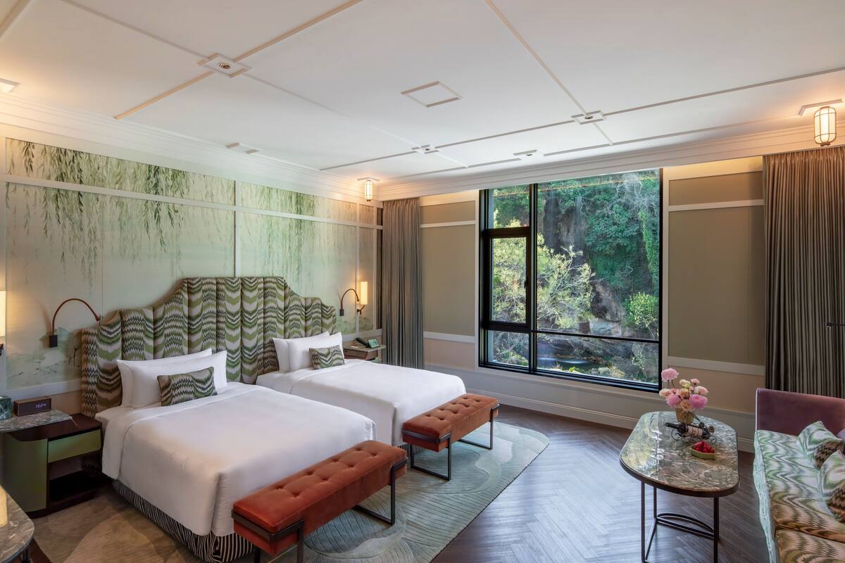 A hotel room with two beds, a sofa, a small table, and a large window with a view of greenery.