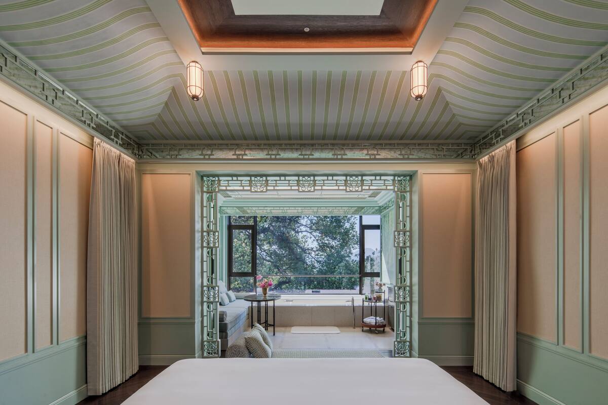 A room with a large bed, a window with a view, a small table with a vase, and a decorative ceiling.