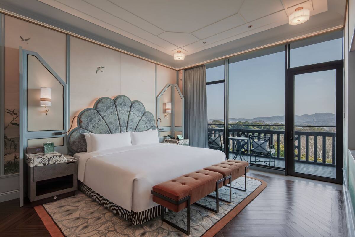 A spacious bedroom with a large bed, a bench, a nightstand, and a view of the outdoors.