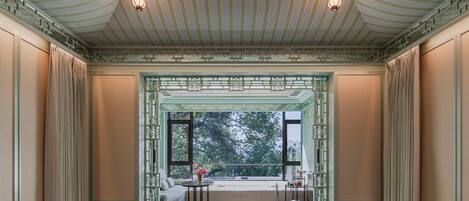 A bedroom with a large bed, a view through a window, and a decorative ceiling.