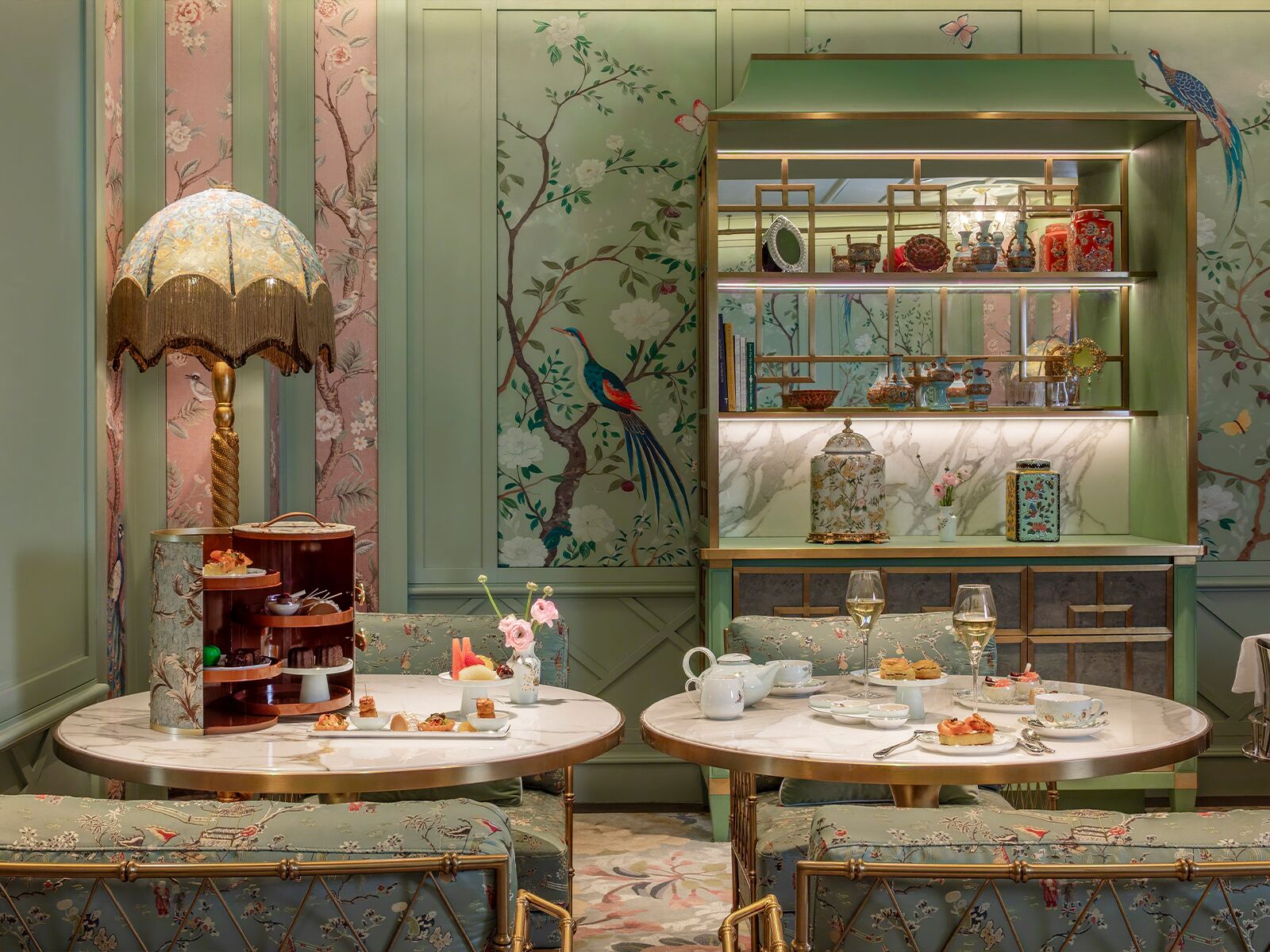A room with two round tables set for tea service, a decorative lamp, and a bookshelf with various items.