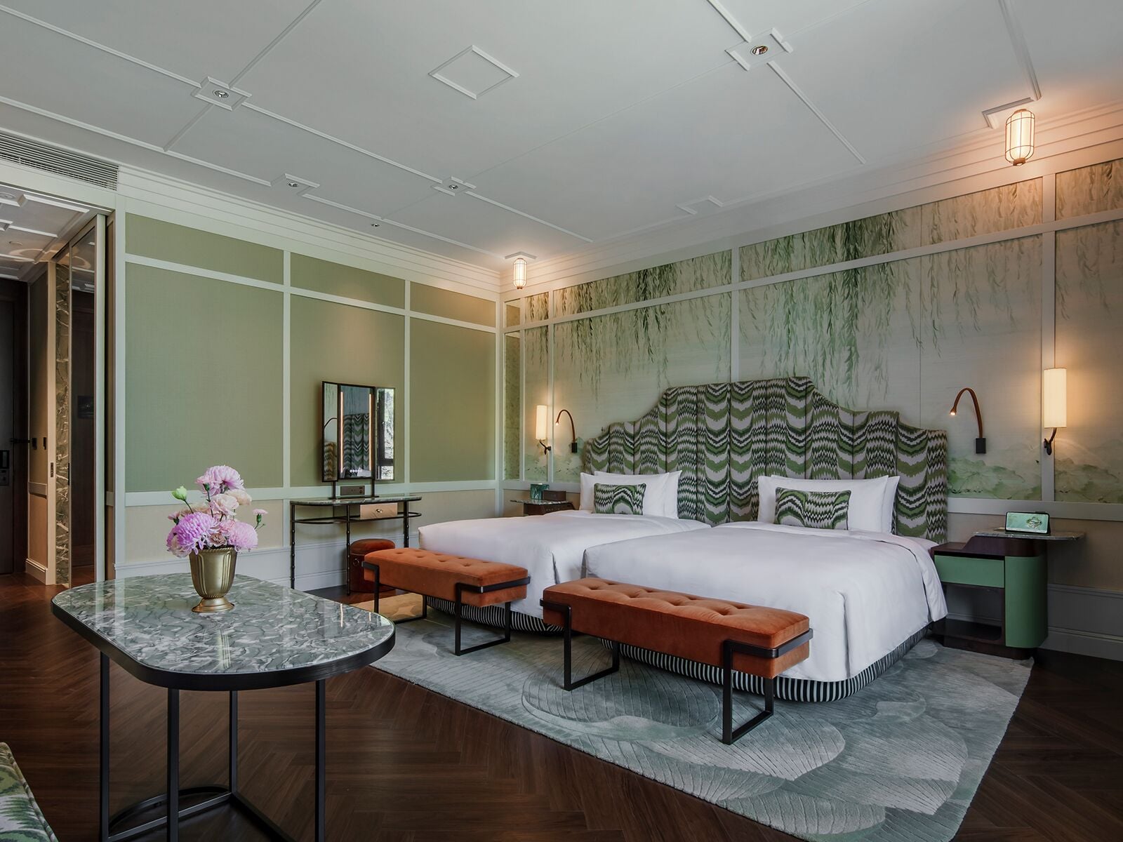 Una habitación de hotel con una cama grande, dos mesitas de noche, una mesita redonda pequeña y un banco.