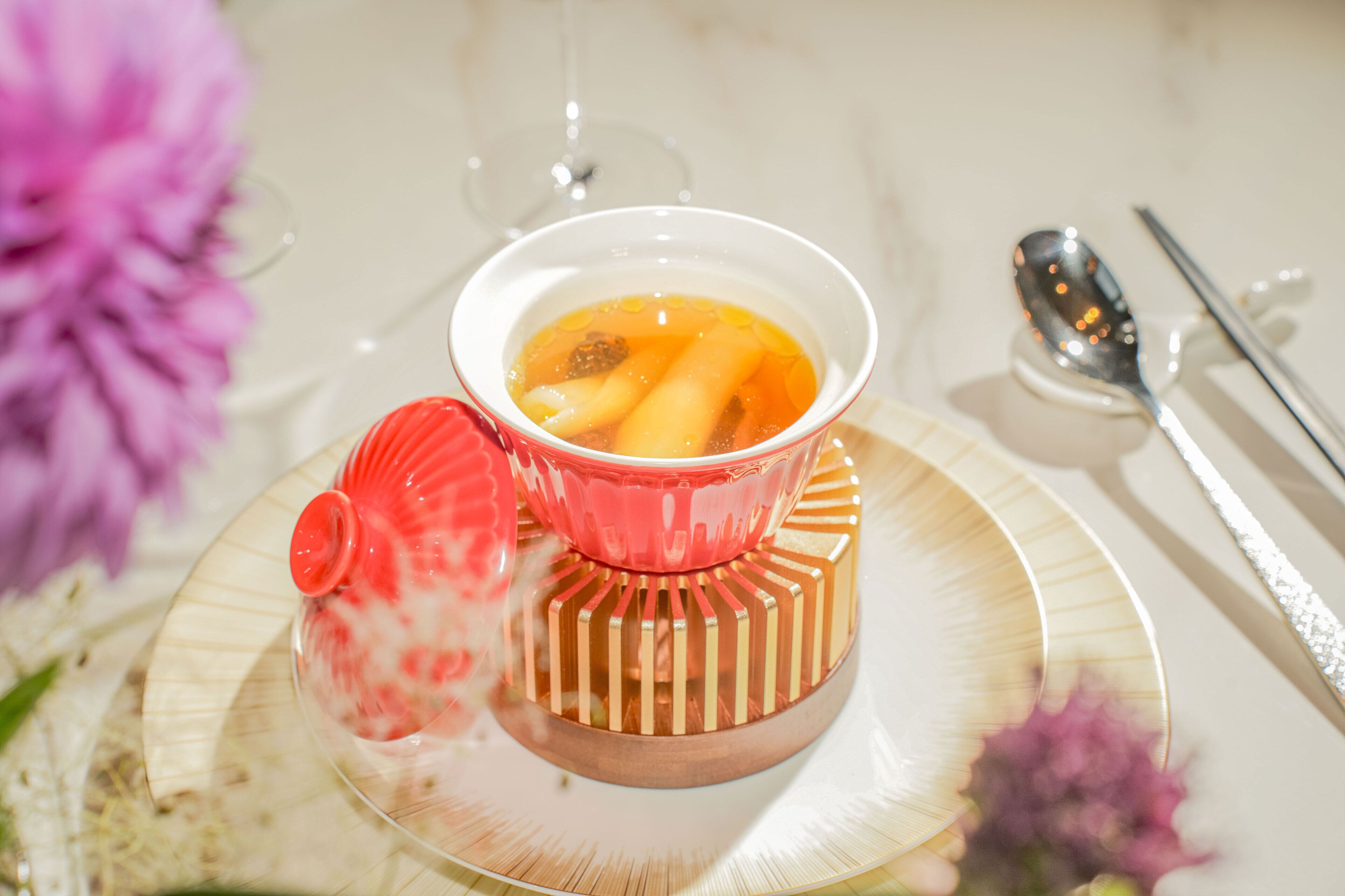 A clear glass bowl with a red lid on a white plate, accompanied by a spoon and a knife on a table.