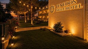 Exterior de un hotel con un letrero que dice 'Tenedos ISkole Hotel', asientos exteriores y luces colgantes.