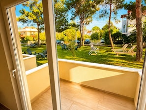 A balcony with a view of a grassy area, trees, and parked cars. - Tenedos İskele Otel (Ezine)