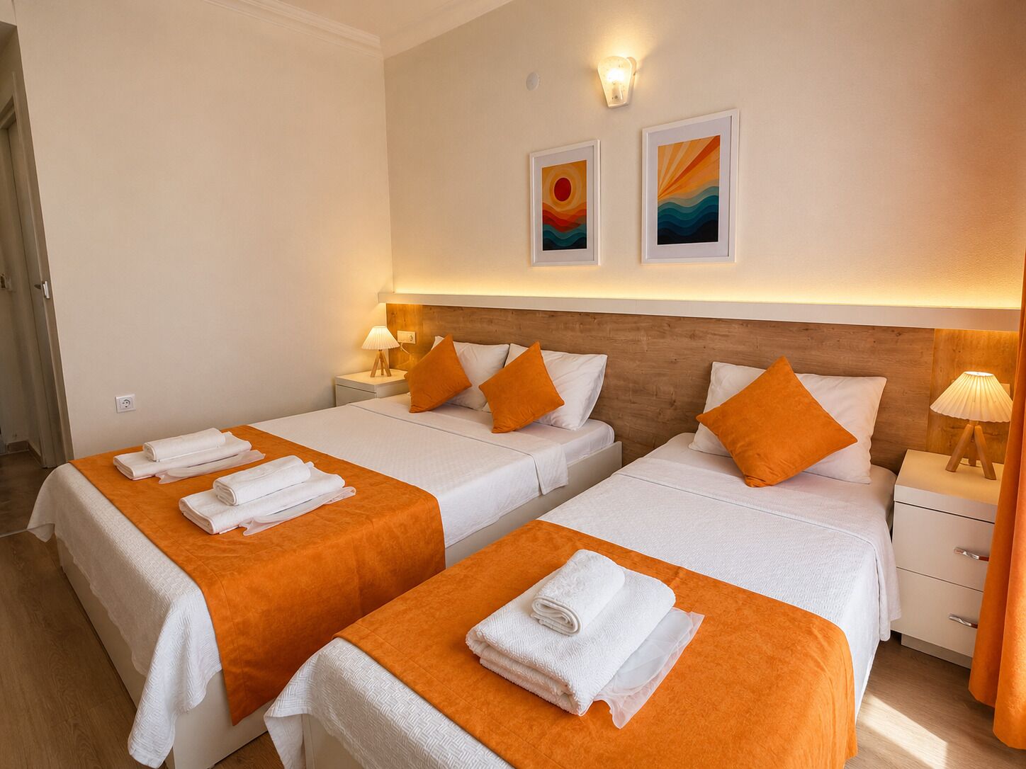 Habitación de hotel con dos camas, cada una con colcha naranja y ropa de cama blanca, cabeceros de madera, mesitas de noche con lámparas y dos lámparas fijadas a la pared.