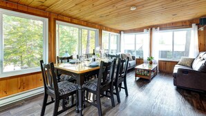 A wooden-paneled room with a dining table set for four, a sofa, and large windows offering a view of trees.