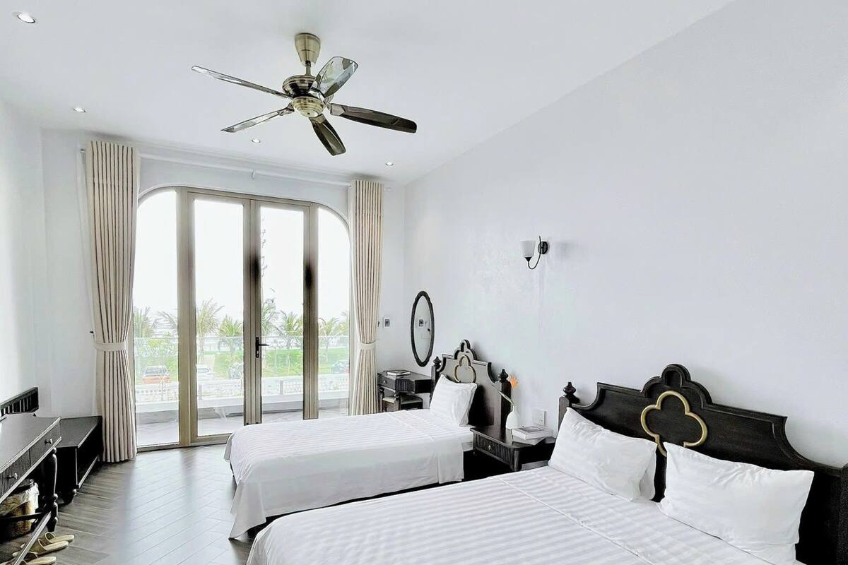 Een hotelkamer met twee bedden, een plafondventilator, een groot raam met gordijnen, een bureau met stoel, en uitzicht op de omgeving.