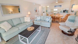 Un salon avec un canapé bleu clair, une table basse en bois foncé et un fauteuil à bascule. On aperçoit à l’arrière-plan un coin repas avec une table et des chaises en bois.