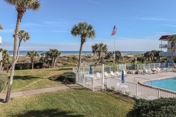 A pool area with lounge chairs and a flag, surrounded by palm trees and a view of the beach.