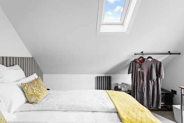 Une chambre moderne avec une lucarne, un lit recouvert d’une literie blanche et d’un plaid jaune, un radiateur et un portant à vêtements avec deux sweats à capuche.