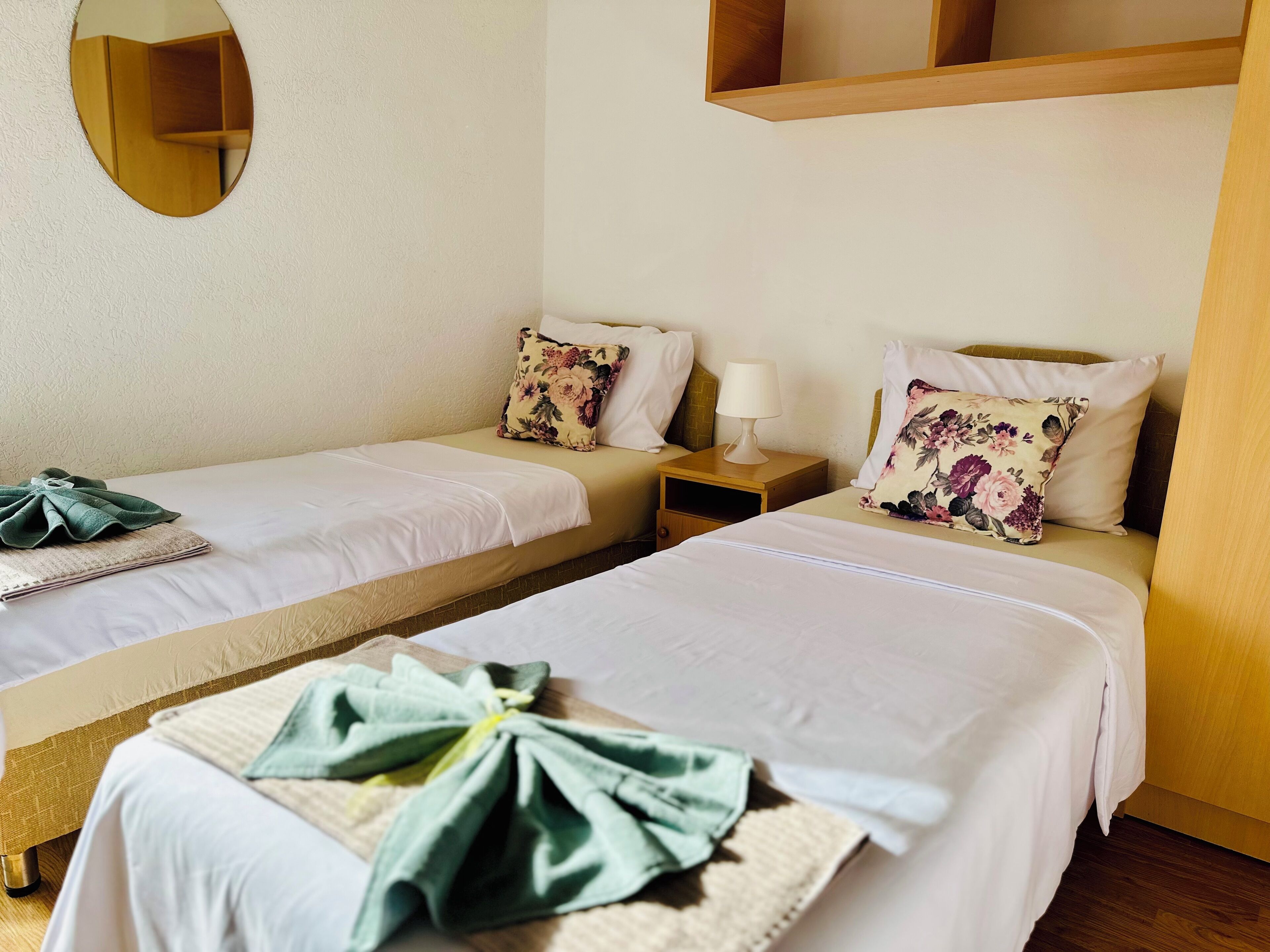 A hotel room with two single beds, a bedside table with a lamp, a mirror on the wall, and a neatly made bed with a towel.