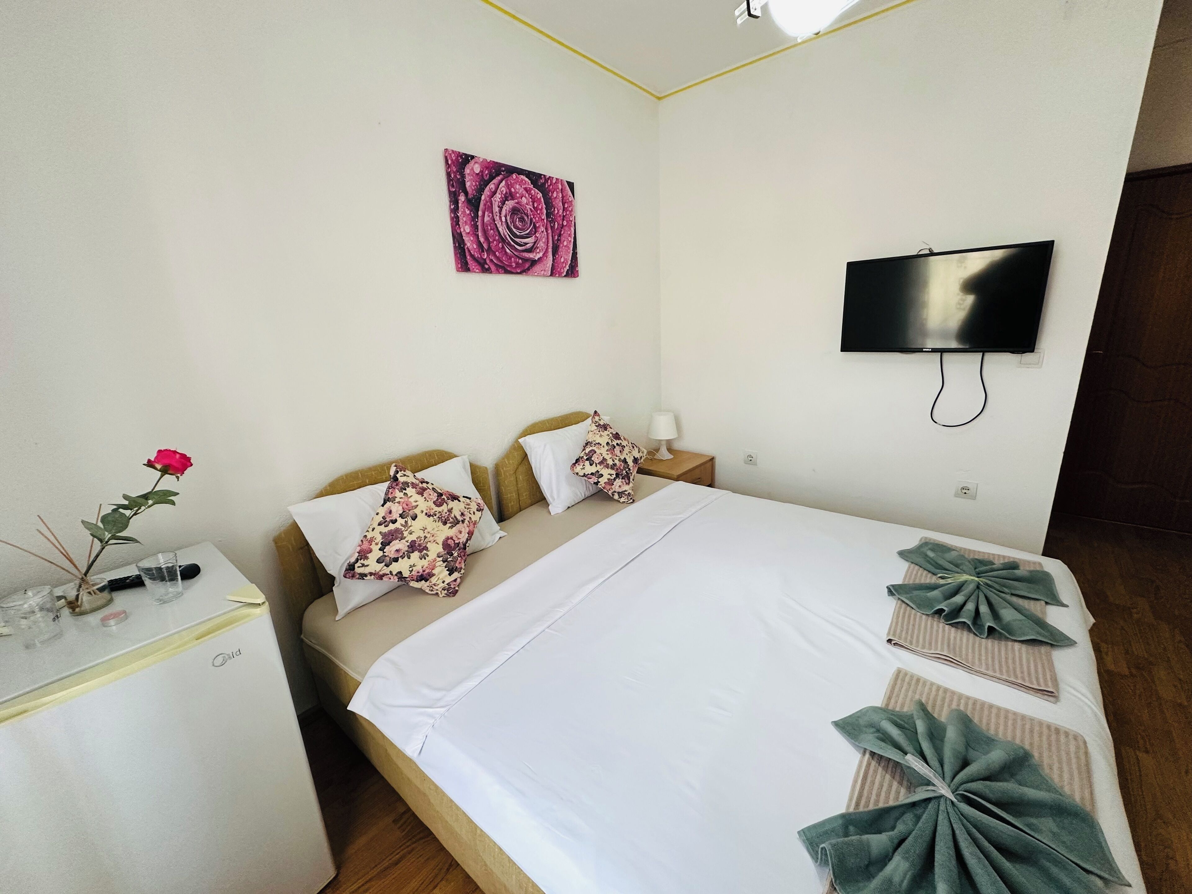 Habitación de hotel con cama, televisor montado en la pared, escritorio pequeño con un jarrón de flores y un cuadro en la pared.