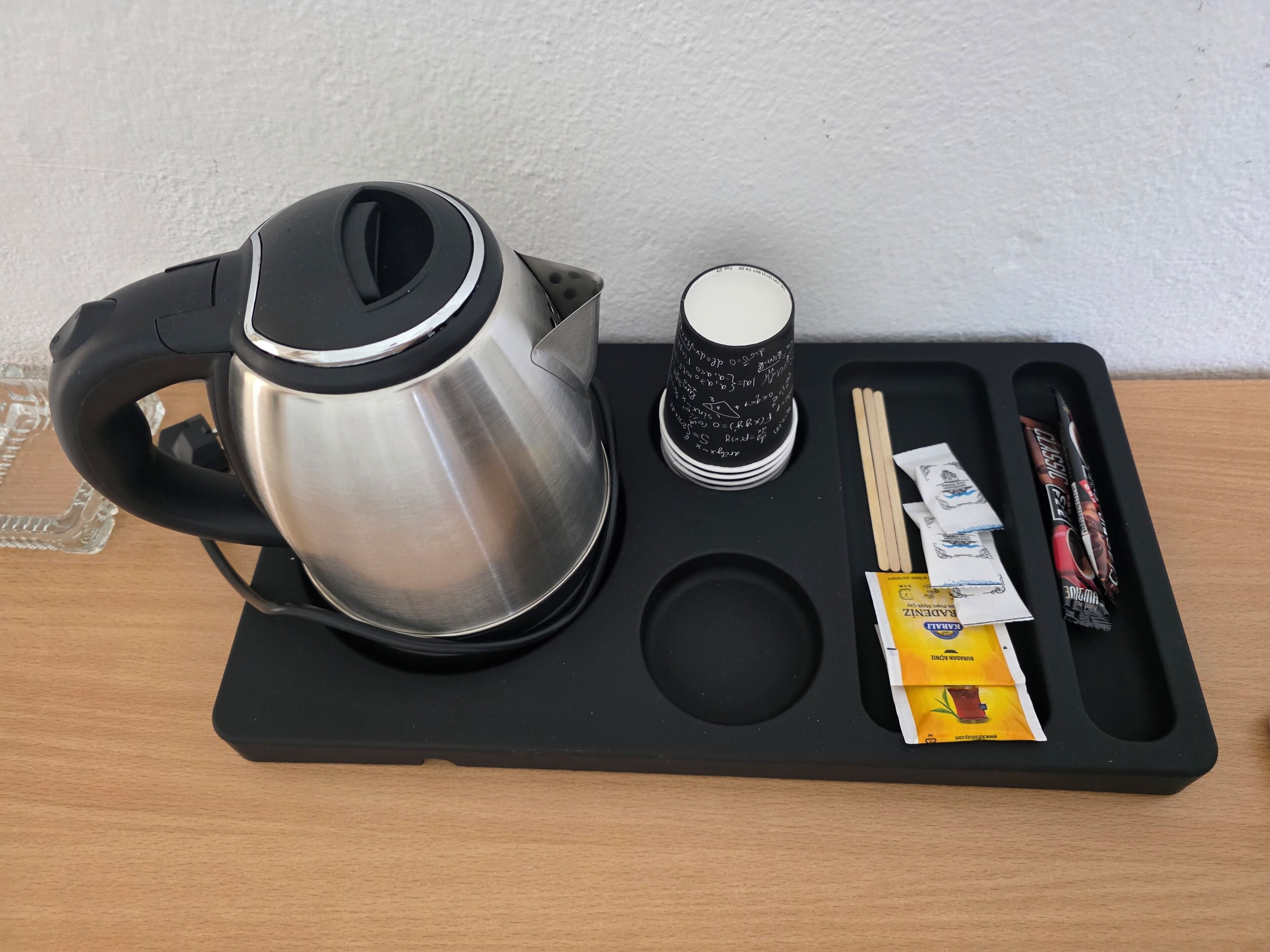 A black tray with a stainless steel kettle, a cup with black text, chopsticks, a pen, and a packet of snacks.
