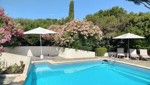 Una piscina rectangular con agua azul cristalina, rodeada por una terraza con baldosas. Hay dos sombrillas blancas que dan sombra, y una fila de sillas de descanso blancas a lo largo del borde de la piscina.