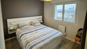 Un dormitorio con cama, mesitas de noche, un ventanal con vistas a edificios y una lámpara en el techo.