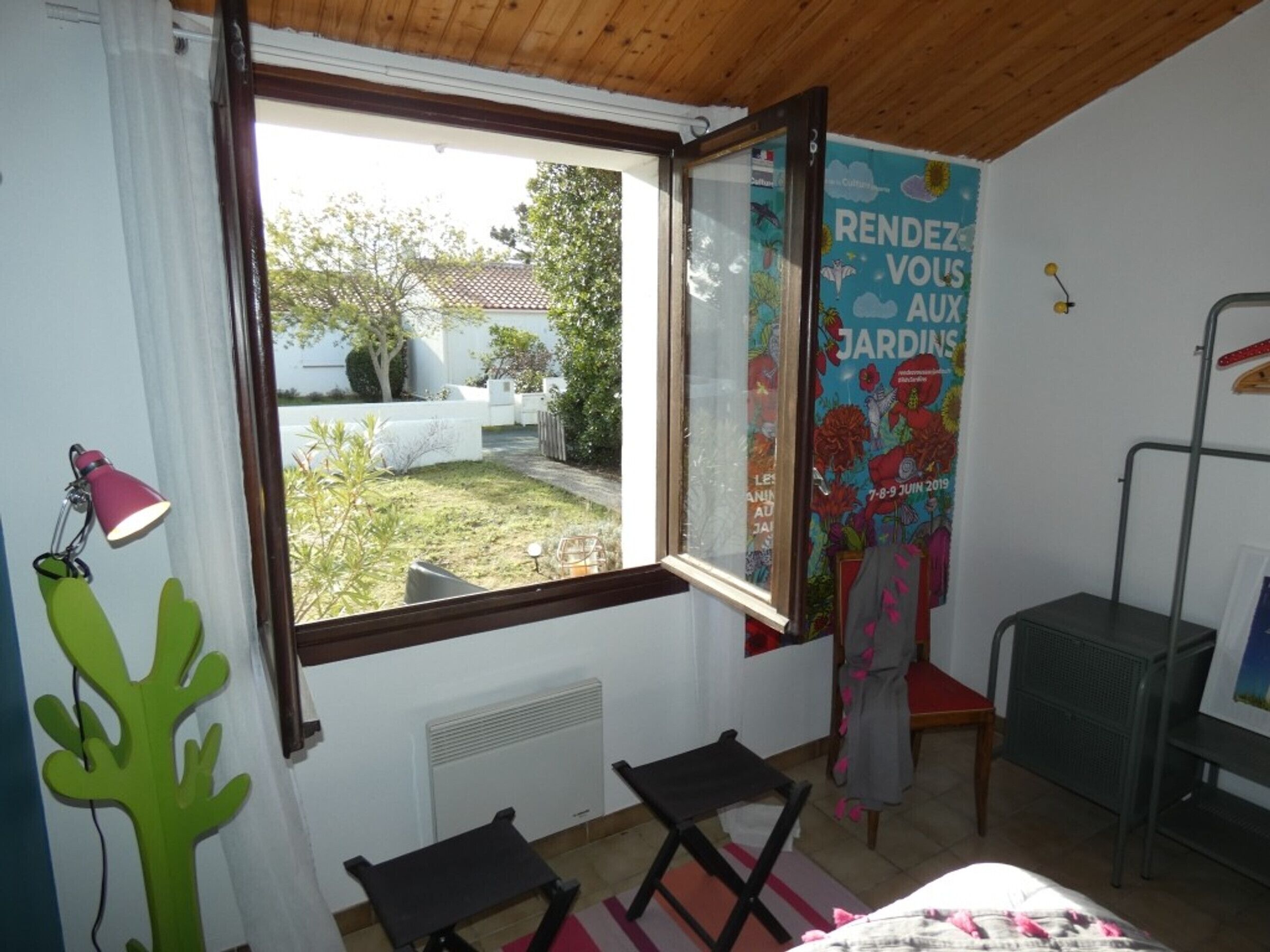Ein Zimmer mit großem Fenster, Heizkörper, kleinem Tisch, Stuhl und einem Poster an der Wand.