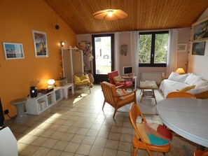 A cozy living room with a wooden ceiling, a round dining table, and a sofa. There are two windows with curtains, a bookshelf, and a lamp.