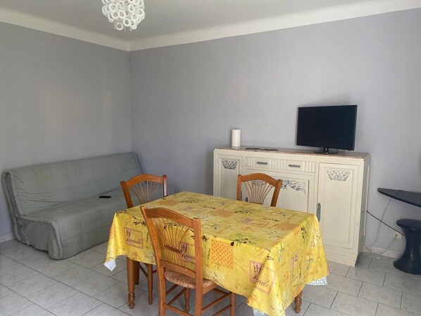 A dining area with a table covered by a yellow tablecloth, two wooden chairs, a grey sofa, and a television on a cabinet.