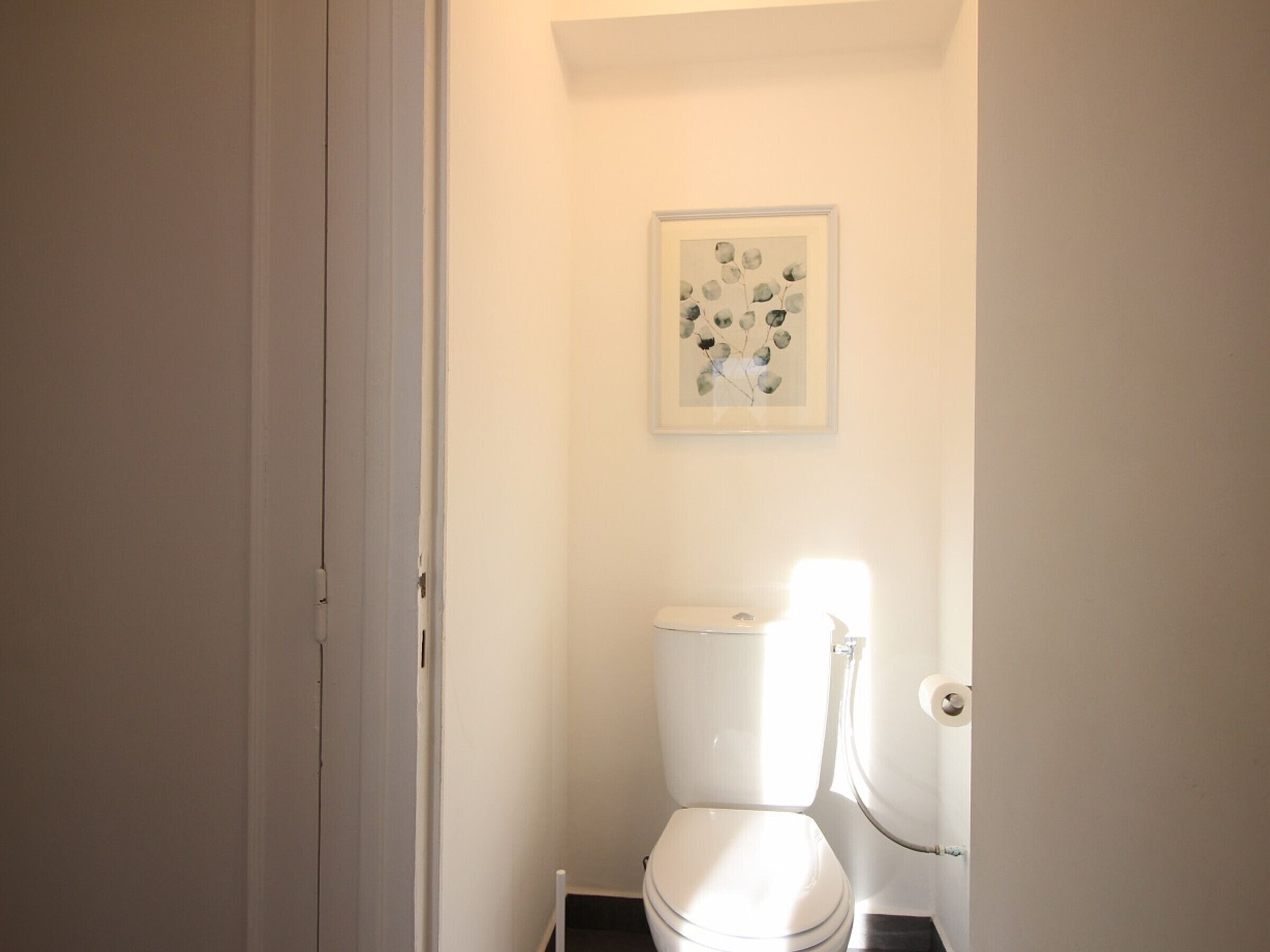 Une petite salle de bain blanche, avec des toilettes, une œuvre d’art encadrée au mur et une porte donnant sur une autre pièce.