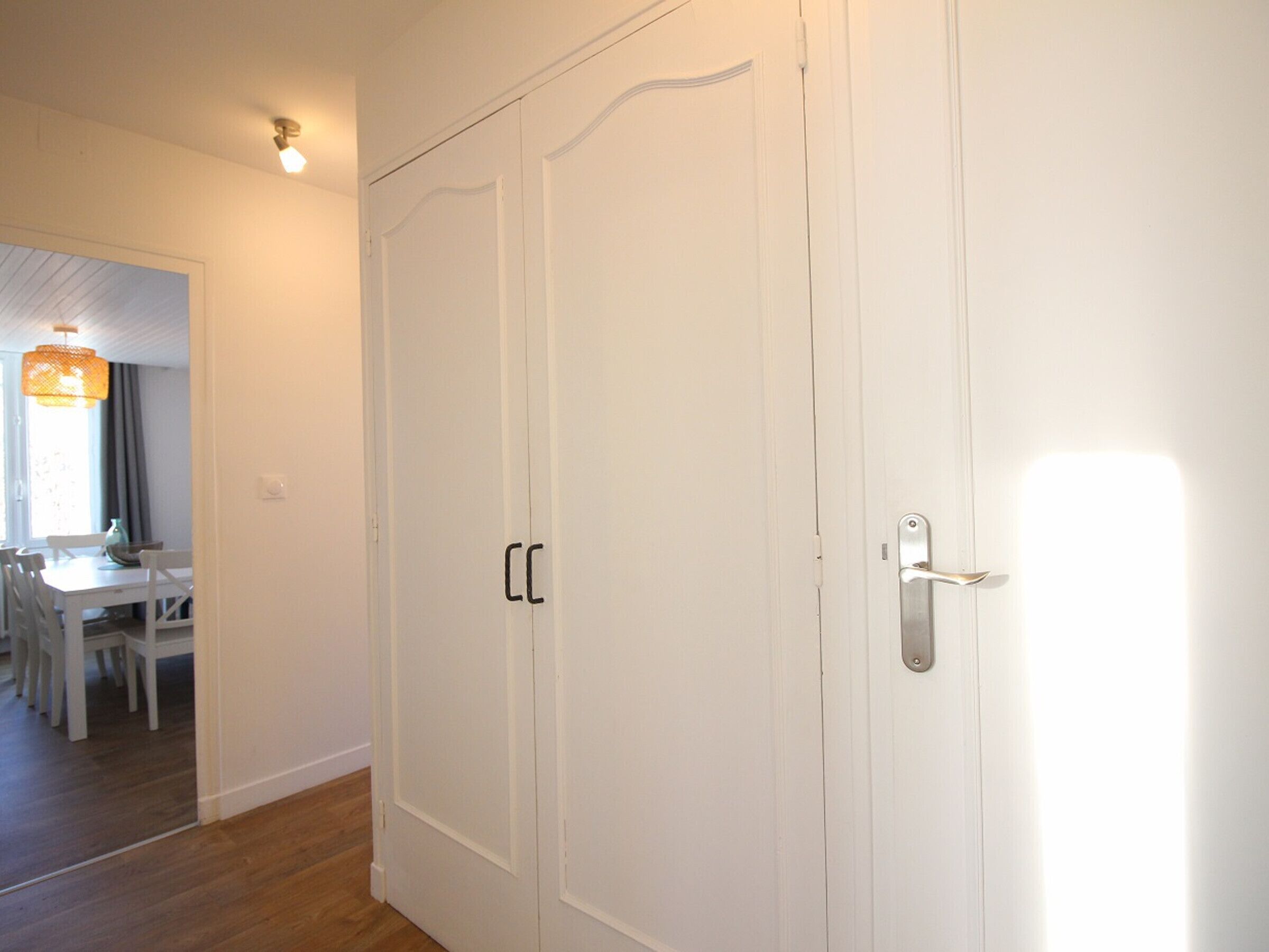 A white wardrobe with two doors and a handle, a dining table with chairs, and a hanging light fixture.