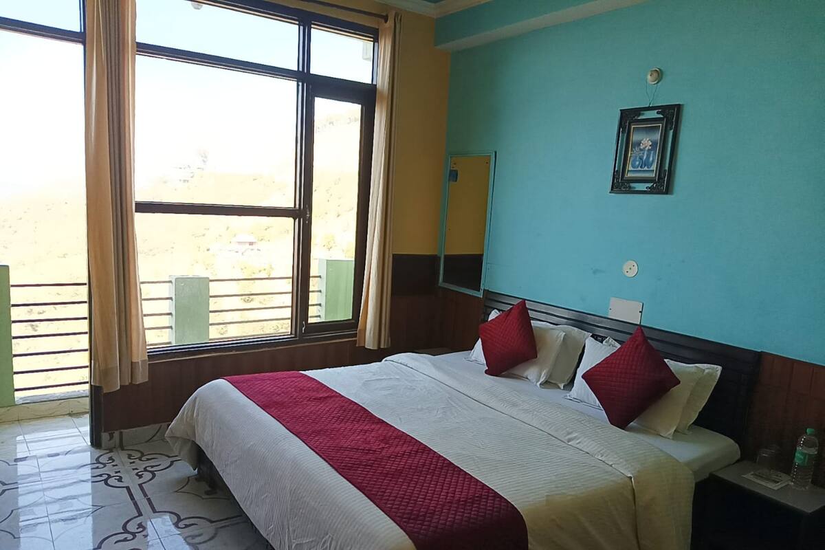 A hotel room with a large bed, a window with curtains, a bedside table with a bottle, and a wall-mounted picture.