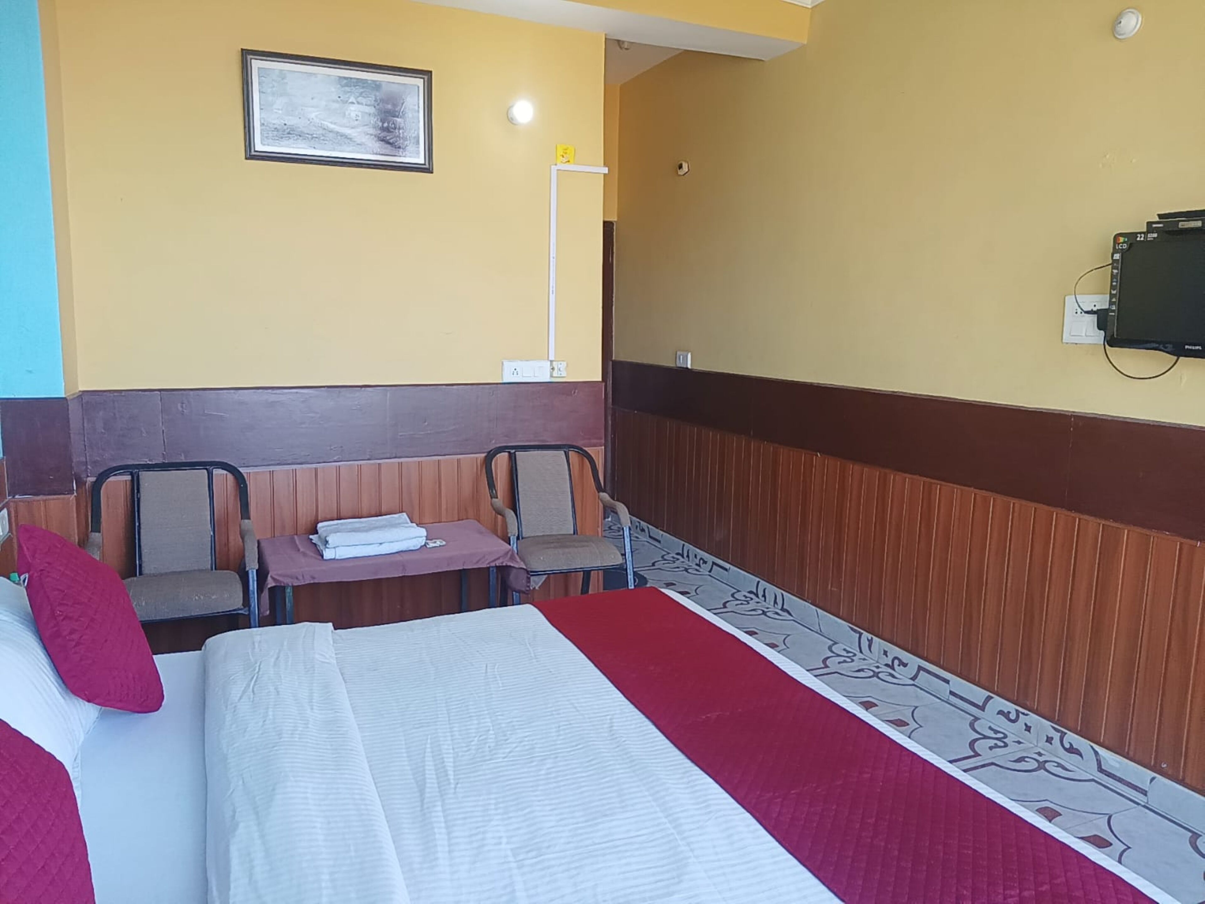A hotel room with a bed, two chairs, a small table with towels, a wall-mounted TV, and a framed picture.