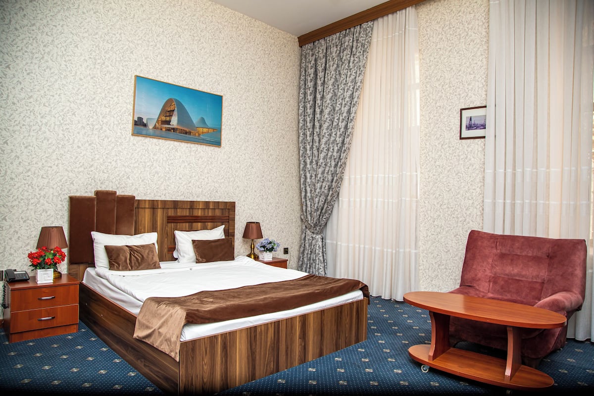 A hotel room with a wooden bed, a red armchair, a small wooden table, a nightstand with a lamp, and a painting on the wall.