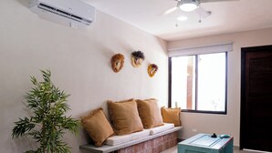 A modern living room with a ceiling fan, air conditioning unit, a sofa with cushions, a small table, a potted plant, and a window with natural light.