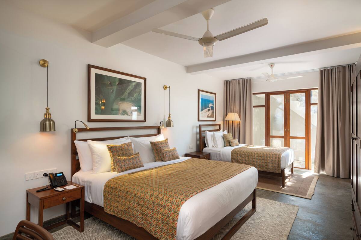 A hotel room with two beds, a ceiling fan, artwork on the walls, and a view through the window.