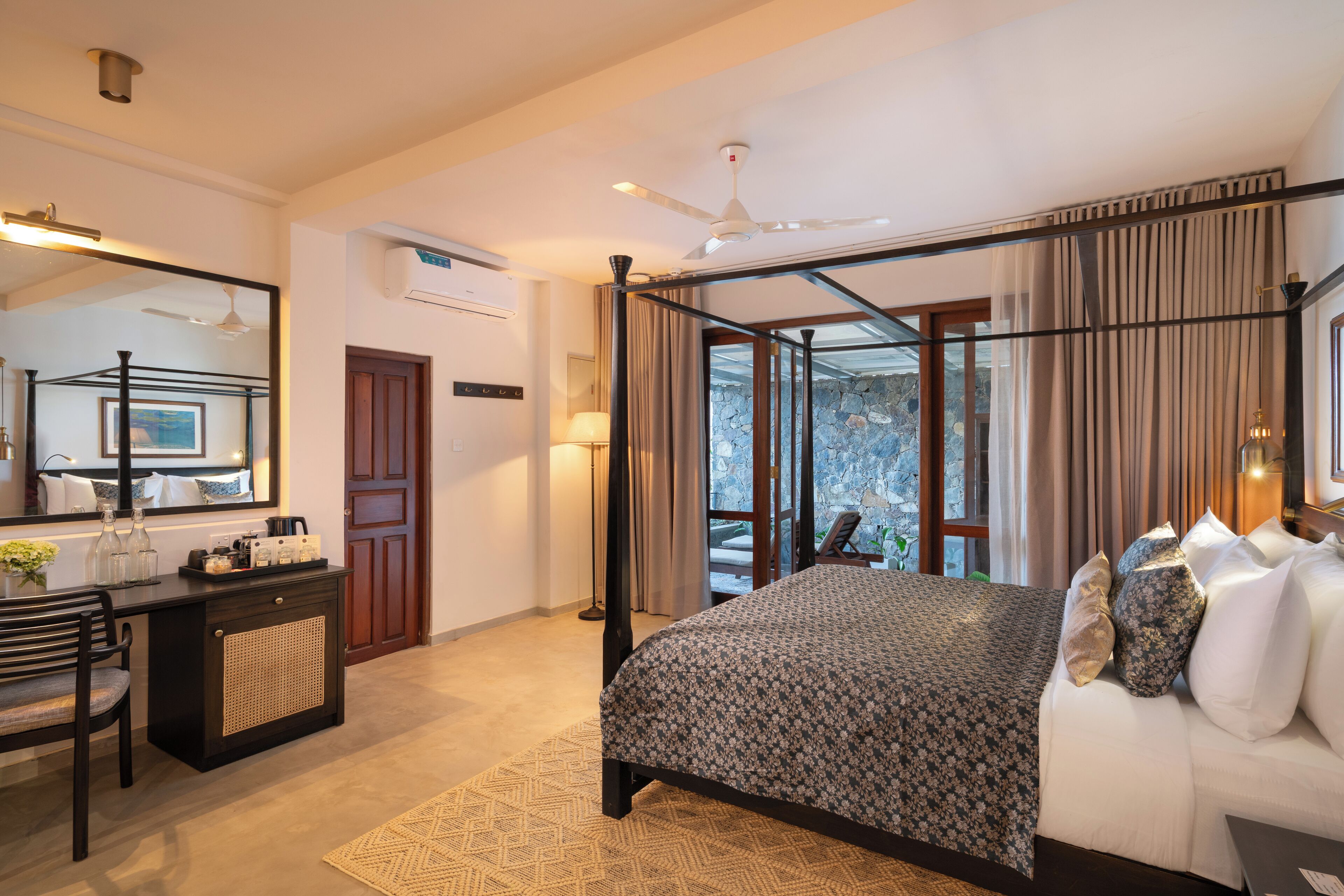 A bedroom with a four-poster bed, a desk with a chair, a mirror, and a view of the outdoors.
