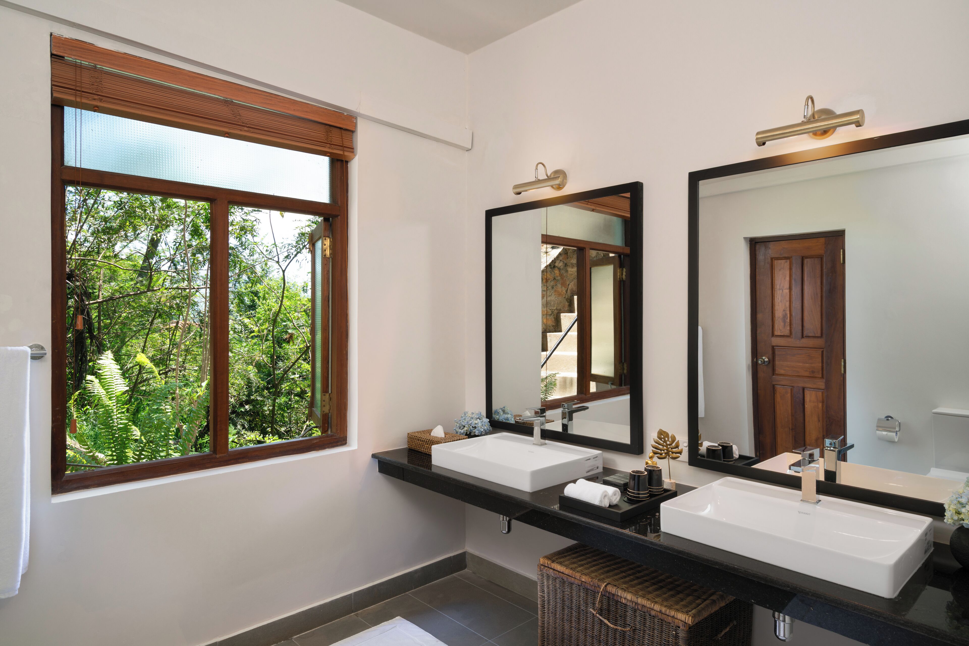 A bathroom with two sinks, a large mirror, and a window with a view of greenery.
