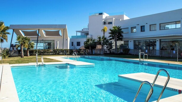 A swimming pool with clear blue water, surrounded by a white building with balconies and palm trees. - Modern 3 bed garden apt next to pool Casa Banderas (La Cala de Mijas)