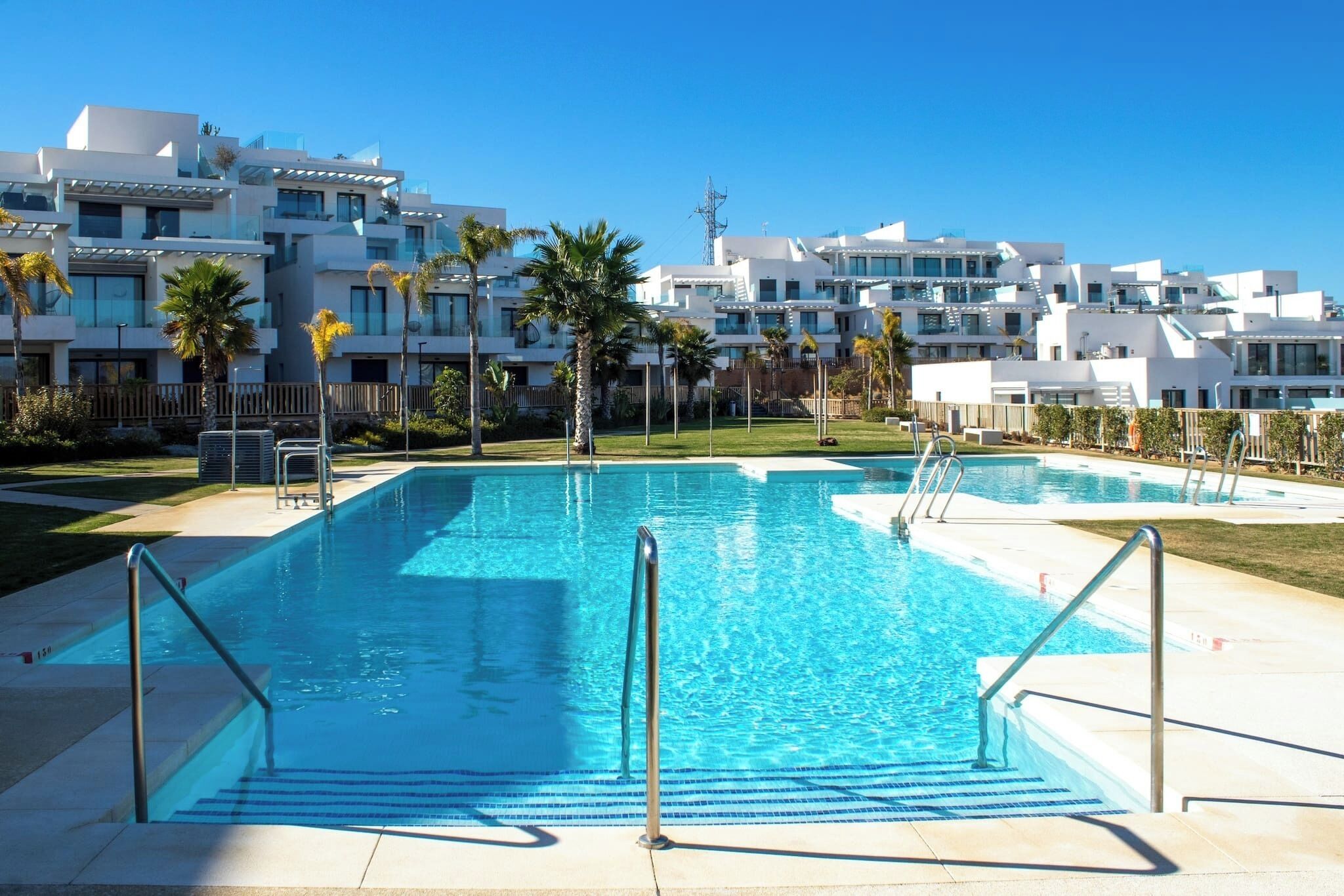 A swimming pool with clear blue water, surrounded by modern white buildings with balconies and palm trees.