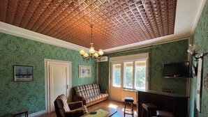 Una sala de estar con papel tapiz estampado, un sofá, sillones, una mesa de centro, una araña y un televisor montado en la pared.