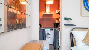 A compact room with a washing machine, a small kitchen area, a bed with a headboard, and a wall-mounted shelf. - Charming Studio moments from Portobello (London)