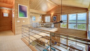 A spacious loft-style room with a high ceiling, wooden walls, a balcony with a railing, and large windows offering a view of the outdoors.