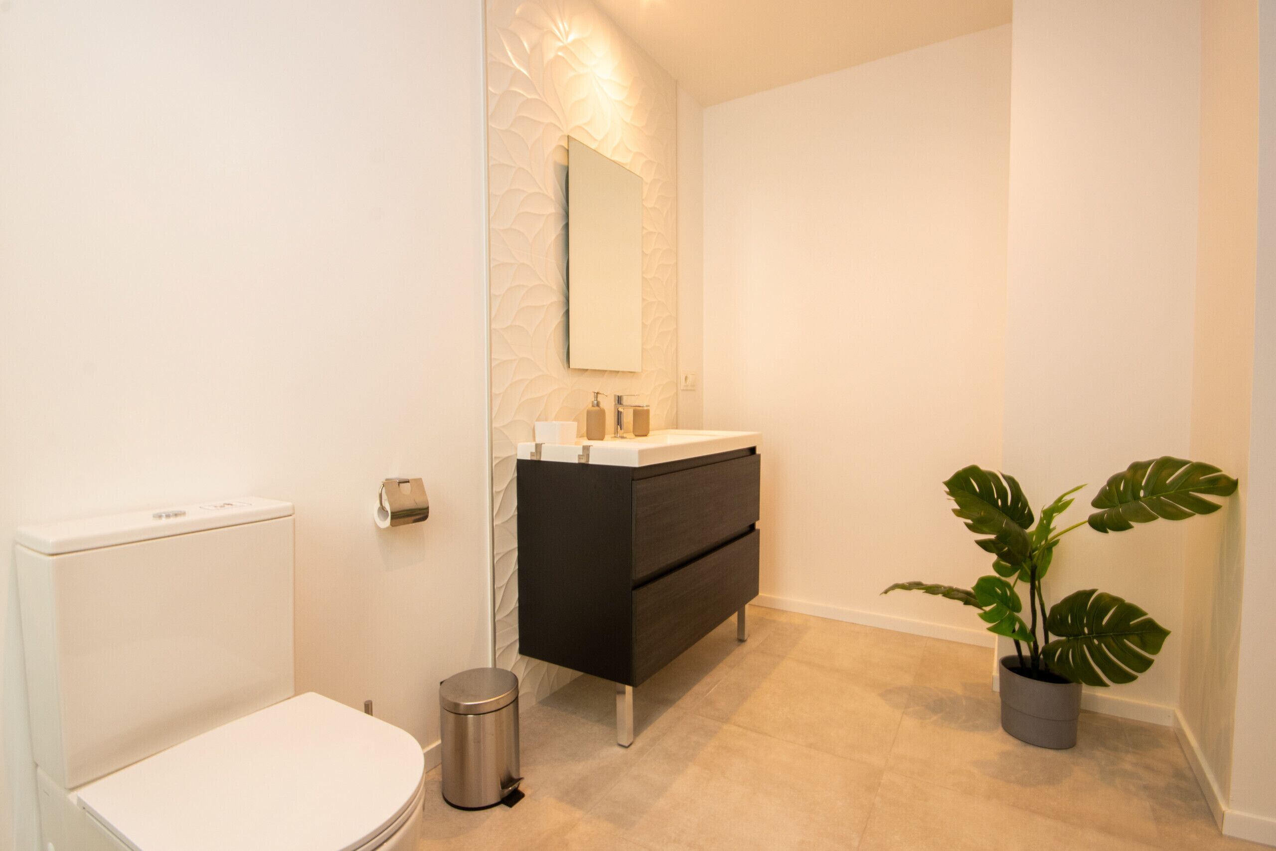 Een moderne badkamer met een wit toilet, een zwart wastafelmeubel, een spiegel en een potplant.