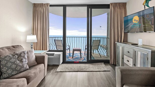 A hotel room with a balcony, a sofa, a TV, and a desk with a lamp. - Palms Resort 1406 - Oceanfront! (Myrtle Beach)