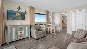 A living room with a sofa, a dining table with chairs, a TV mounted on the wall, and a view of the beach through the window. - Palms Resort 1406 - Oceanfront! (Myrtle Beach)