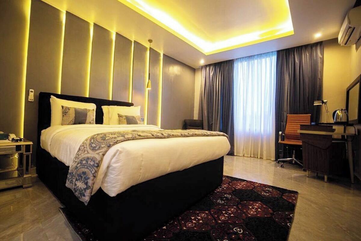 A modern hotel room with a large bed, bedside tables, a desk, a chair, and a large window with curtains.
