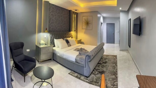 A modern bedroom with a large bed, a nightstand, a chair, and a small table. - Ted G Hotel (Port Harcourt)
