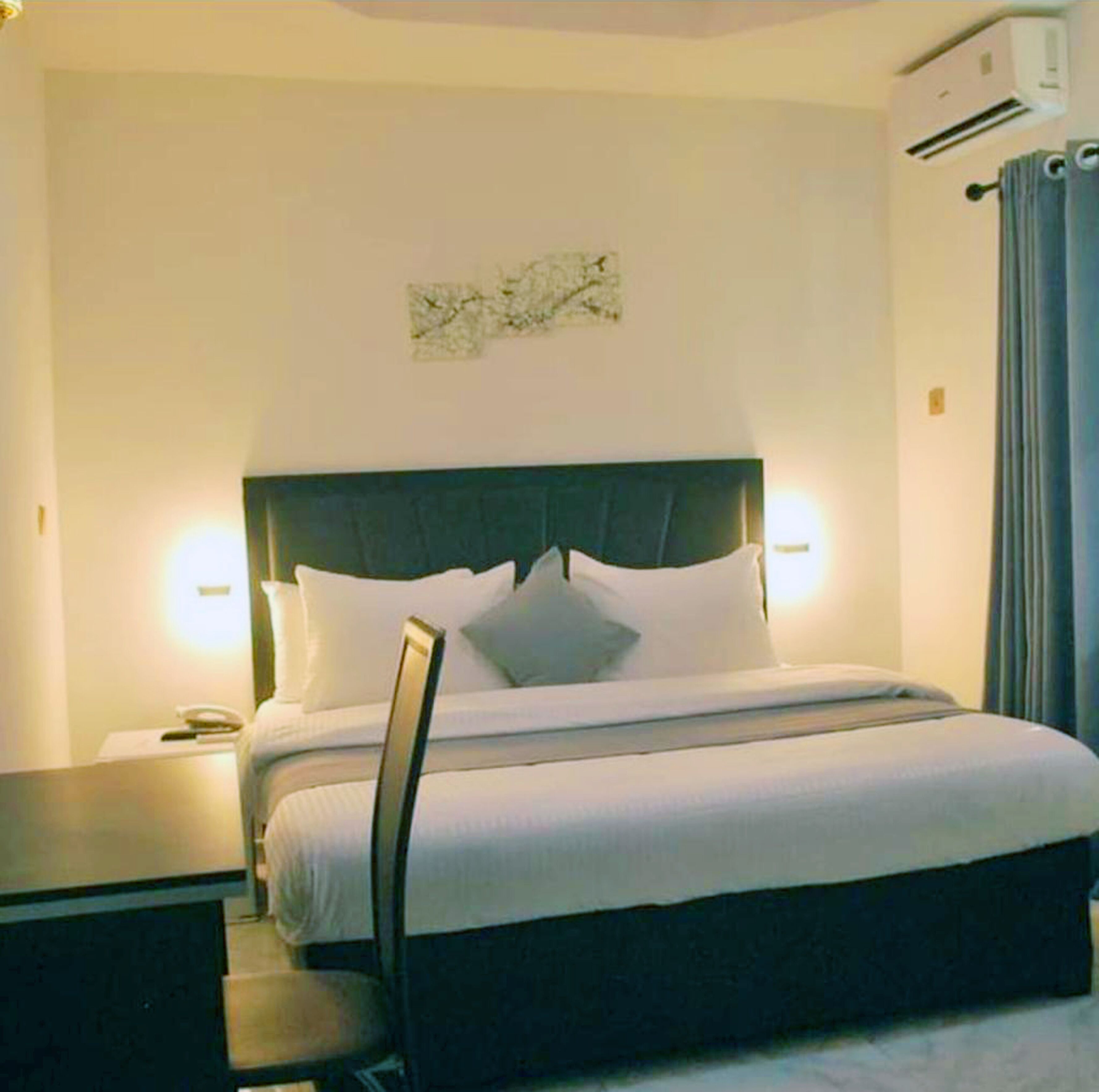 Una habitación de hotel con una cama grande, una mesita de noche, una lámpara y un aire acondicionado.