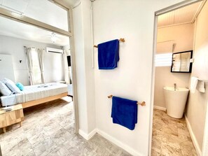 A hotel room with a bed, a bathroom with a sink, and two blue towels hanging on the wall. - Eden On The Sea (Bridgetown)