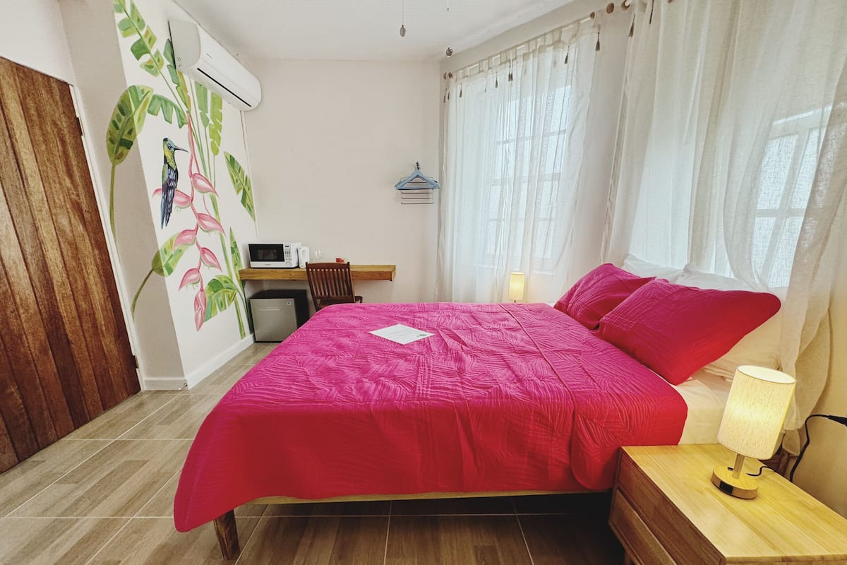 A bedroom with a bed, bedside table, lamp, and a desk with a microwave and books.